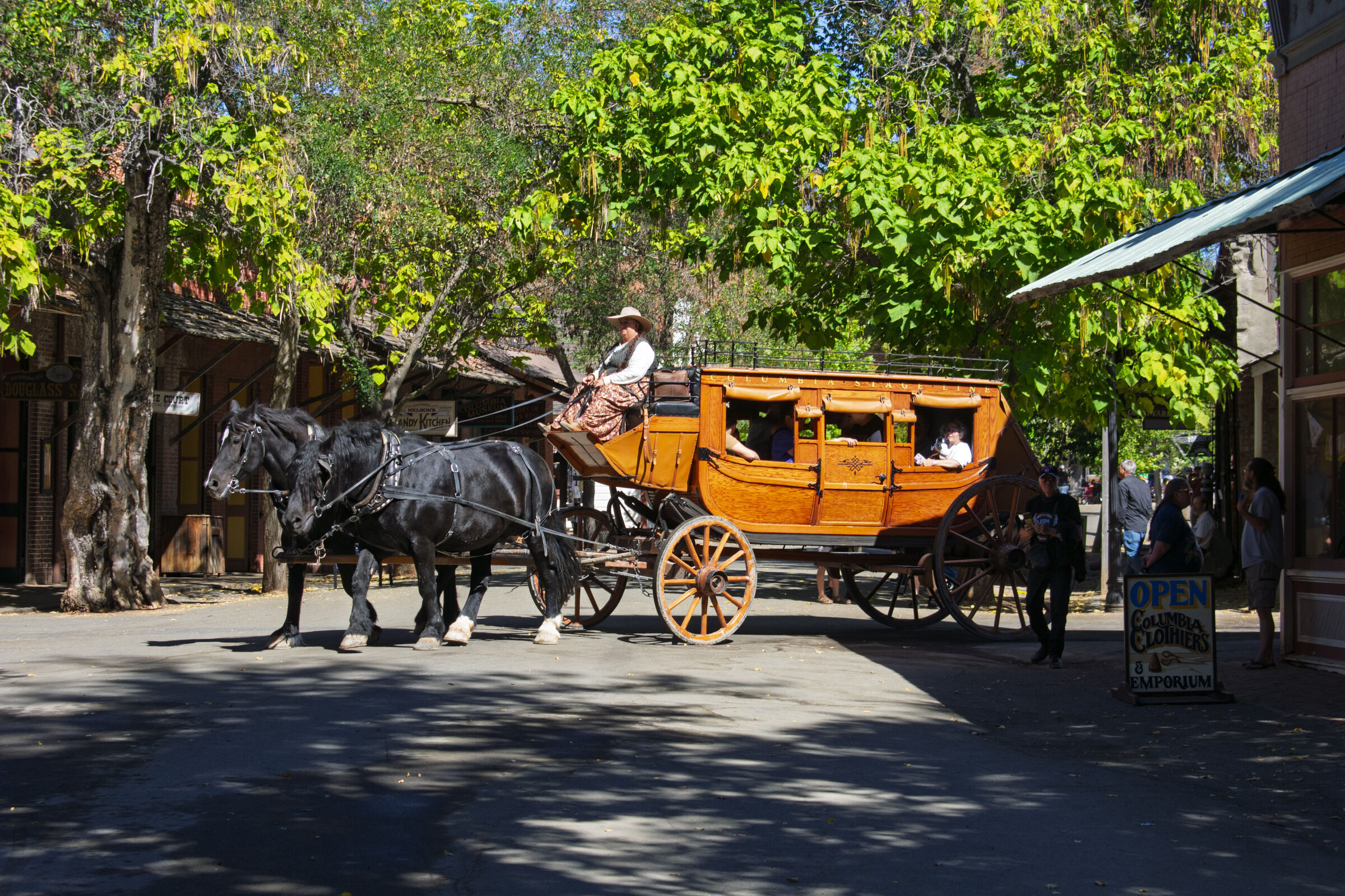 A team of horses pulling an orange stagecoach down a shaded street in Columbia State Historic Park
